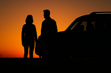 Image shows the silhouettes of a man and a woman standing at the front end of an SUV.
