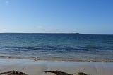 View from a sandy beach across a narrow stretch of calm sea to a distant headland.