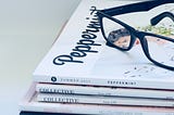 A stack of glossy magazines with a pair of glasses resting on top