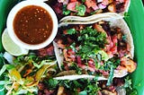 An overhead shot of a platter of soft tacos, with a ramekin of sauce and a lime wedge, on a green plate.