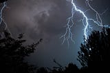 A navy blue sky with clouds, silhouetted by tropical trees. But there is an electric blue lightning that strikes the sky