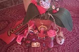 A potted orchid on a red patterned rug surrounded by small objects, including a Reese’s Peanut Butter Cups pencil case, a red Chinese tassel, a blue-and-white bird figurine, a ladybug decoration, several pins, a small doll, a pink rose ornament, a miniature bouquet, a stuffed bird, and other small keepsakes arranged around the base of the plant.