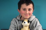 A young boy around 9 or 10 years old. He has brown hair and a small fringe. He’s smiling and sitting against a blue wall. He’s holding a stuffed animal of a meerkat. His cheeks are a little rosy, and is also wearing a grey sweater.