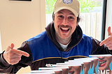 attractive 50-ish year old man in beige hat points at books and smiles.