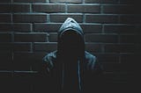 Image of a hooded man, face not visible, stood against a brick wall, beneath a light