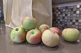 A small pile of reddish green apples sits next to a bag filled with even more apples.