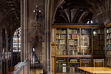 A neo-Gothic style library. Upstairs with a hallway and arched walls and pillars. On the right side, a book-case of rare books and a wooden table on the bottom right with two chairs.