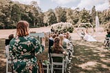 Photo of me in a green floral dress, behind the seated guests. During outdoor wedding ceremony. Standing, painting.