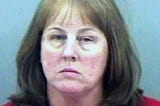 In a grainy mugshot, a middle-aged woman frowns at the camera. She has light brown hair. and is standing against a light blue wall.