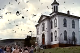 Color still from the film, “The Birds,” showing children and actress Tippi Hedren fleeing from birds swarming in the sky with a building in the background.