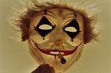 A mask of a crazy clown face and wild hair with a cigarette sticking out of its mouth is held up by a hand, partially in view
