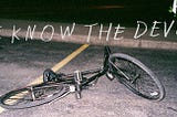 A bike lies on its side in a dark parking lot. Chalky text superimposed above it reads: WE KNOW THE DEVIL.