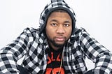 Adrian Prince seated with his elbows resting on his raised knees as he looks directly into the camera in a close-up head shot. Adrian Prince is wearing a black and white plaid pullovers with a gold necklace laying atop the black t-shirt with red writing that he’s wearing beneath his plaid pullover.