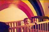 Baby me, Jenny Lane, standing in my wooden crib, hands holding the edge, painted on the wall of my room is a purple first rainbow