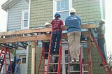 People with construction helmets stand on ladders, constructing a porch roof in front of a nearly completed Habitat for Humanity house. One is the author’s younger daughter. (Photo by author.)