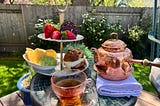 A tiered tea tray with a egg and cheese sandwich on a sourdough pretzel bun, corn chips and guacamole, and strawberries and dark chocolate on the top tier. Next to it is a pink glass cup filled with black tea and a copper teapot. It all sits on a small garden table outside.