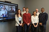 A group photo of nine people smiling. Three people are smiling on a tv screen from a virtual platform.