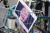 Photograph of a wire basket of a bicycle containing a to-go cup of coffee and a protest placard showing Donald Trump shouting with a reddened face with “Fight Tyranny” written in red at the top. (Photo by Samantha Sophia for Unsplash.)