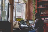 Description: A woman sitting in front of her table working on her laptop while facing the window. Object: person  Action: working on the computer Context: working