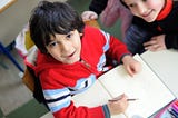 A small boy is sitting in front of a notebook with a pencil in his hand. He looks up at the camera with a smile. The girl next to him smiles at him admiringly.