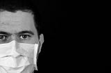 Black and white image of a person wearing a cloth medical mask over nose and mouth, set against a black background