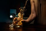 Against a dark background, a woman in a sleeveless, tan top slams a glass of whiskey down hard on a mahogany table, sloshing liquid everywhere.