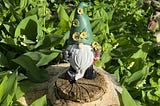 A garden gnome with a green hat decorated with yellow flowers stands atop a tree stump, surrounded by vibrant greenery. The gnome has a long white beard, black boots, and holds a small watering can in one hand and a flower pot in the other. Sunlight highlights the lush foliage and the intricate details of the gnome’s features.