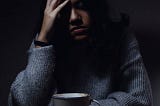 Depressed woman sitting in a dark room. Her hand rests on her forehead in obvious stress. A cup of coffee sits in from of her. She is wearing a sweater.