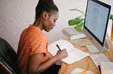 An African girl sitting down and writing into her notepad in front of a Mac computer screen