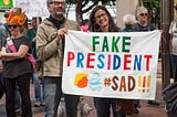 Rally attendees hold a banner containing a cartoon profile of Trump and  colorful letters in the style of a tweet, reading “FAKE PRESIDENT #SAD!!!”
