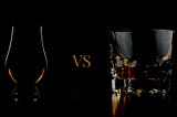 A Glencairn glass with liquor on the left hand side and a tumbler with liquor on the right hand side with the letters ‘VS’ between them against a black background