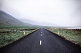 A two-lane road heads toward large hills and a cloudy sky.