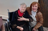 Sister Emily Meisel in wheelchair with author Judith Valente kneeling by her side.