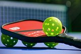 Pickleball paddle and balls, sitting on a pickleball court.