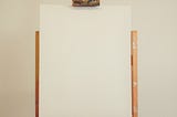 A wooden easel with a blank white canvas stands against a cream wall.