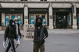 Activist of color stands outside Westminster Abbey holding a sign reading “The social contract was broken”