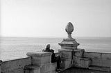 Monochrome photo of an older man sitting on a cement pier staring at the ocean