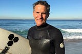 Man in a wetsuit holding a surf board in front of the ocean on a sunny day
