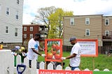 Two black men wearing baseball caps and volunteer t-shirts install signage for a community garden within a neighborhood lot.