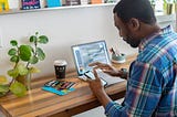 A man sitting at a wooden desk in front of a laptop, looking down at the phone he’s holding in his hand.