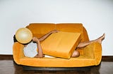 A person stretched out on a mustard colored couch with a balloon covering the face