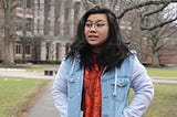 Me, a 20 year old Asian American woman with round glasses, in a sweater and a hoodie outside on my college academic quad.