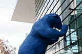 Large outside blue bear sculpture peering into the glass of the front of the Denver convention center. There are trees and building in the background.