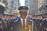 A young man with dark brown skin wears a United States Air Force uniform and stands in the streets of New York City flanked by fellow airmen for the Veterans Day Parade in 2018.