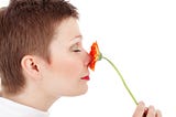 The side view of a short-haired woman smelling a pink flower held up to her nose.