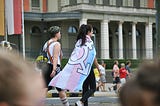 Two people walking along street, one with transgender flag being worn as a cape.