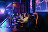 A panel of gamers in headsets look at gaming monitors while intently playing, with their gameplay projected onto large screens behind them.