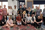 A photo of 18 people, some standing, some sitting, smiling at the camera. Some are holding instruments including lute, sitar, cello and recorder