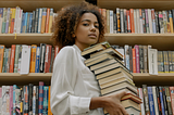 A woman standing in front of a bookshelf carrying a stack of books.