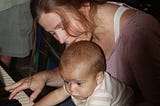 Photo of a mother and very young son (9 months) at the piano together. Her hair is sandy brown, pulled back, with strands falling across her face and down toward the piano. Her son’s eyes bulge out as he grasps for the instrument. One of her hands holds him. The other touches the keys. Both of the baby’s hands are on the white keys.